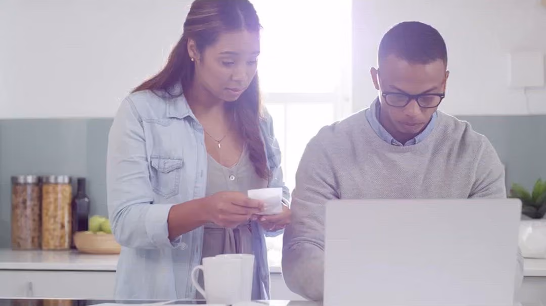 A young couple at their kitchen counter, looking over their finances on a laptop.