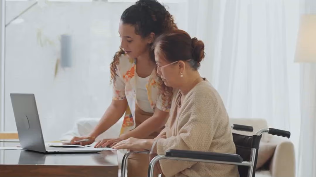 A woman helping an older woman in a wheelchair with documents at a laptop.